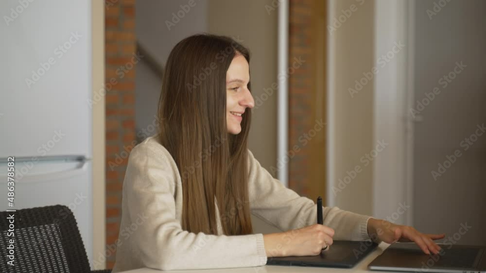 Young brunette retoucher smiles and starts video chat when drawing on pen tablet. Female illustrator holds stylus in hand, answers video call, waves with hand in greeting. Light calm pastel colors. Young brunette retoucher smiles and starts video chat when drawing on pen tablet. Female illustrator holds stylus in hand, answers video call, waves with hand in greeting. Light calm pastel colors.