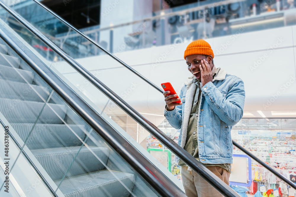 Emotional happy African-American guy in stylish outfit looks at mobile phone going up escalator in shopping mall close view