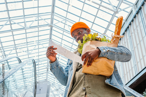 Surprised African-American man in denim jacket looks at receipt total in sales check holding paper bag with products in mall photo