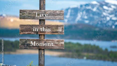 live in this moment text on wooden signpost outdoors in landscape scenery during blue hour. Sunset light, lake and snow capped mountains in the back. photo