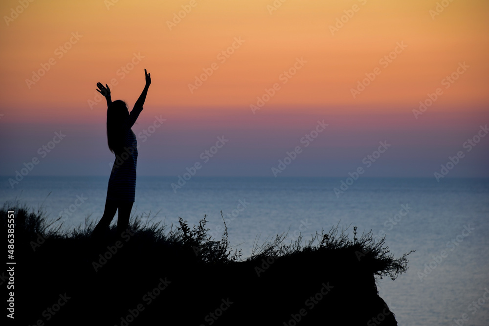 Silhouette of an unrecognizable woman with outstretched arms against the background of the evening sea and sunset
