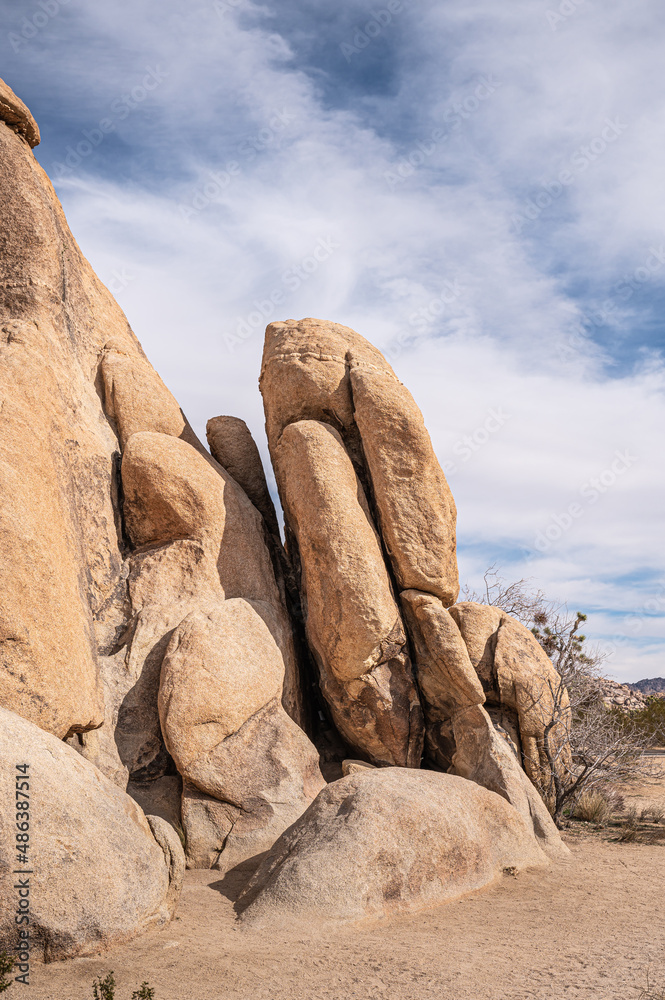 Joshua Tree National Park, CA, USA - January 31, 2022: Closeup of cracked-up, split and beige part of giant boulder on sandy desert floor under blue cloudscape. Bushes in back. Joshua Tree National Park, CA, USA - January 31, 2022: Closeup of cracked-up, split and beige part of giant boulder on sandy desert floor under blue cloudscape. Bushes in back.