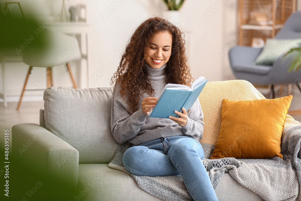 Young African-American woman reading book on sofa at home