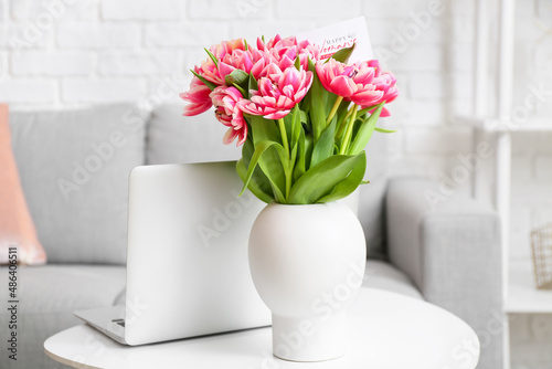 Modern laptop, vase with bouquet of tulips and greeting card on table. International Women's Day celebration