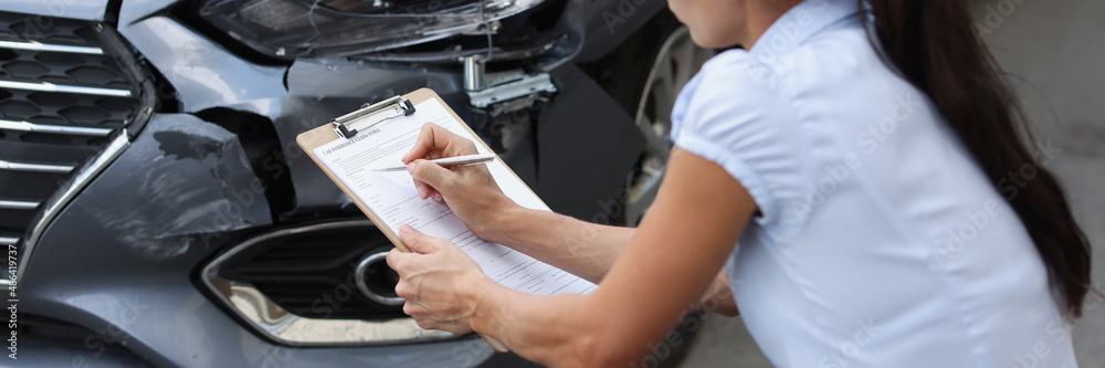 Young woman agent sitting near broken car and writing information into documents