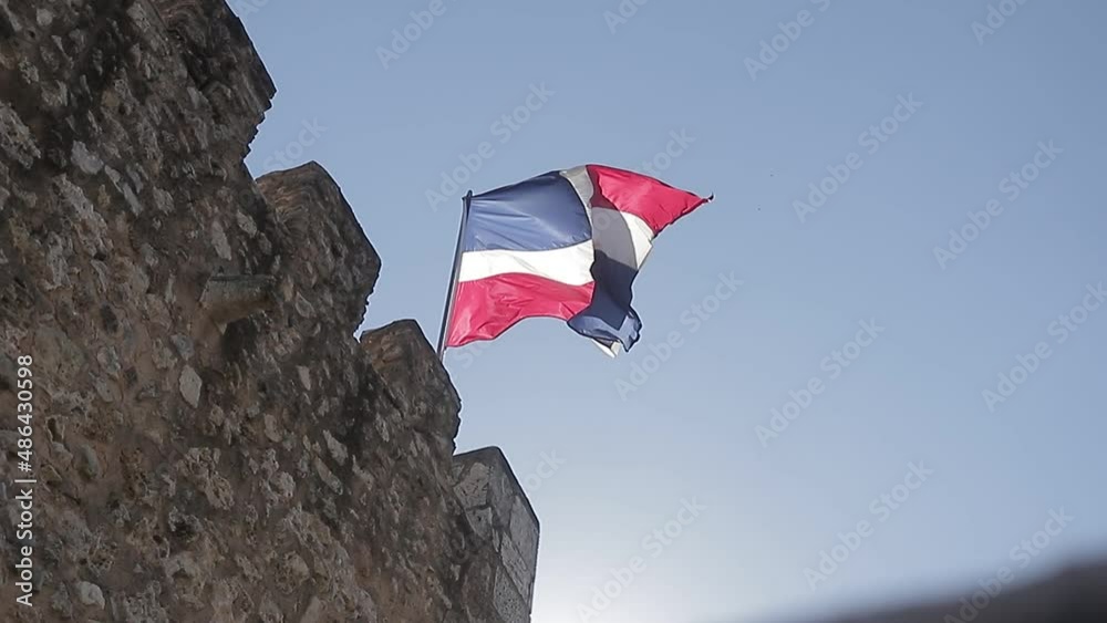 Captura a cámara lenta de la bandera de la república dominicana ondeando en lo alto con un sol radiante detrás y un cielo azul claro.