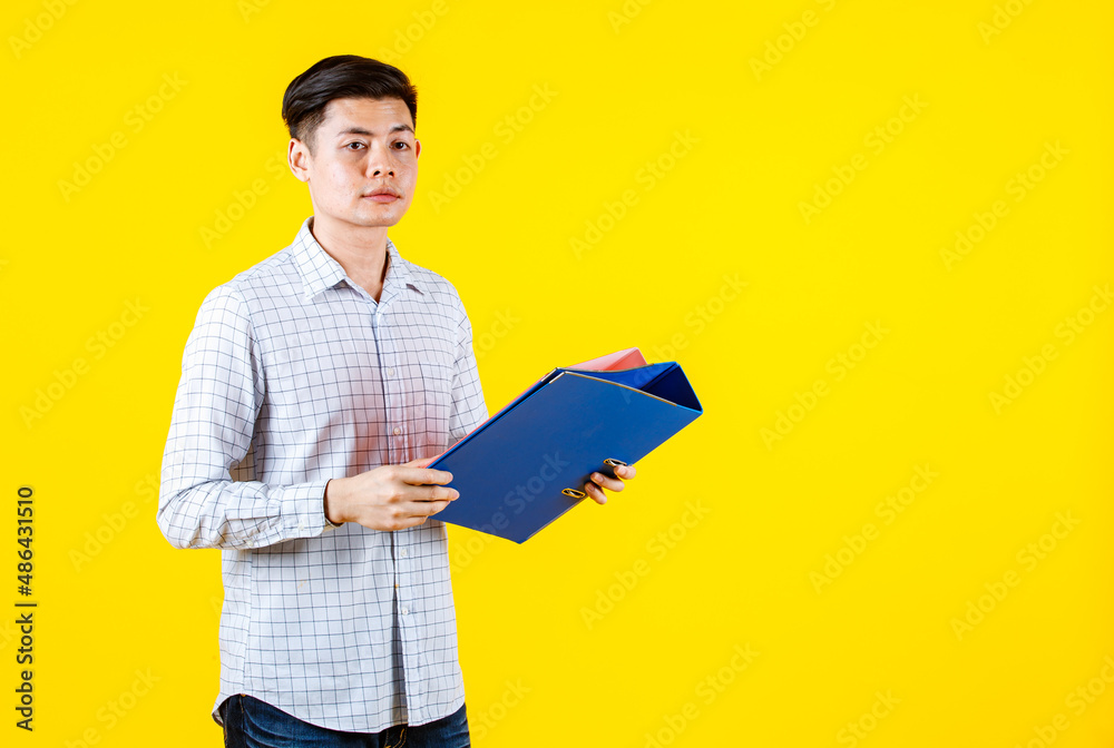 Portrait studio shot of Asian young male businessman employee model in casual outfit standing look at camera holding paperwork document file colorful hard case folders in arm on yellow background Portrait studio shot of Asian young male businessman employee model in casual outfit standing look at camera holding paperwork document file colorful hard case folders in arm on yellow background