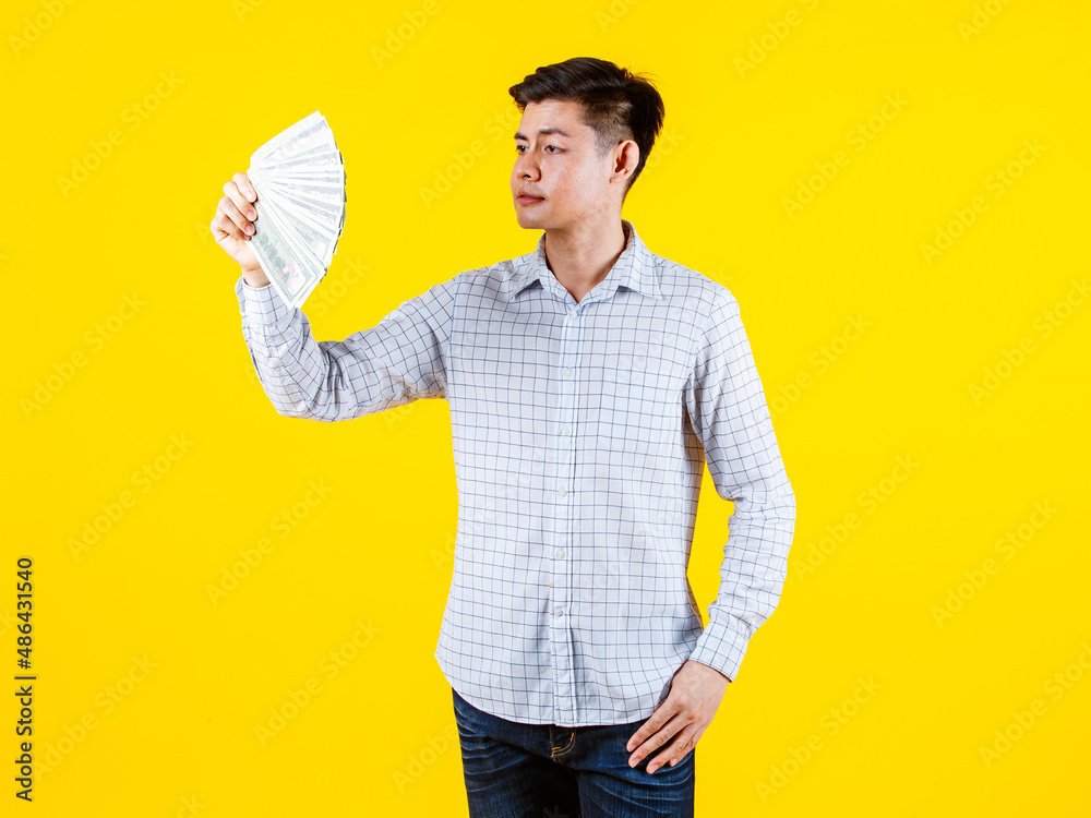 Portrait studio shot of Asian young rich wealthy male millionaire businessman in casual outfit standing smiling look at camera holding stack of hundred dollar banknotes money fan on yellow background Portrait studio shot of Asian young rich wealthy male millionaire businessman in casual outfit standing smiling look at camera holding stack of hundred dollar banknotes money fan on yellow background