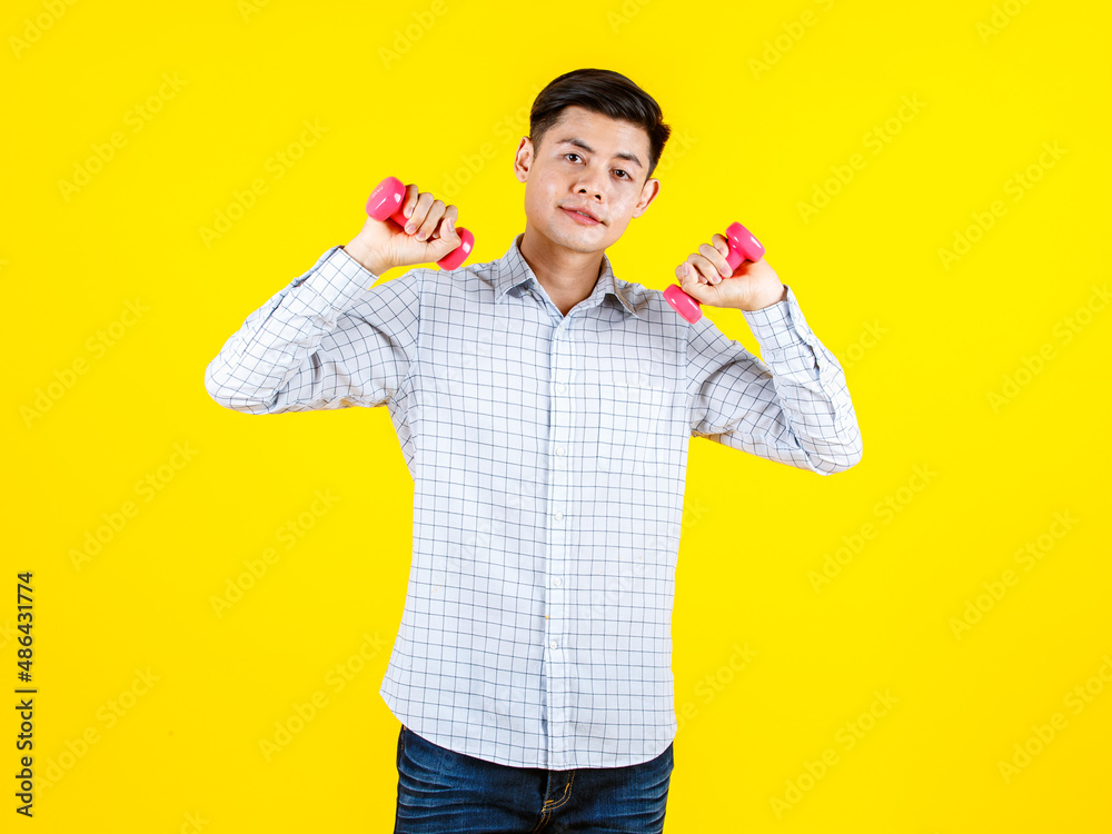 Portrait studio shot of millennial Asian young handsome male sporty athlete model in casual outfit standing look at camera holding two small pink dumbbells lifting exercising on yellow background Portrait studio shot of millennial Asian young handsome male sporty athlete model in casual outfit standing look at camera holding two small pink dumbbells lifting exercising on yellow background