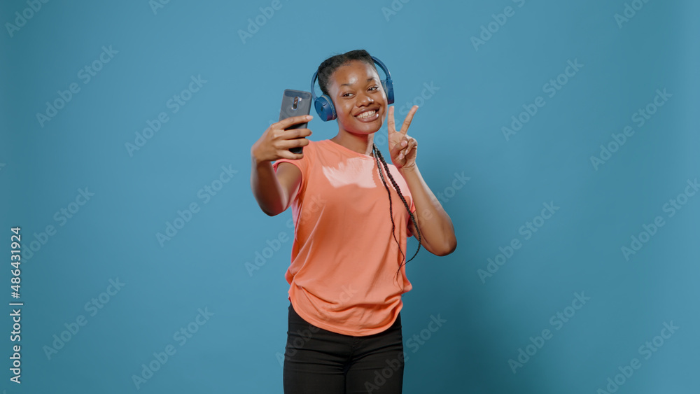 Naklejka premium Modern woman recording dance moves and singing song on headphones in studio. Young adult smiling and holding smartphone to film video on camera. Person using headset and technology.
