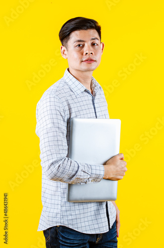 Portrait studio shot of millennial Asian young male businessman in casual outfit standing smiling holding laptop computer on yellow background