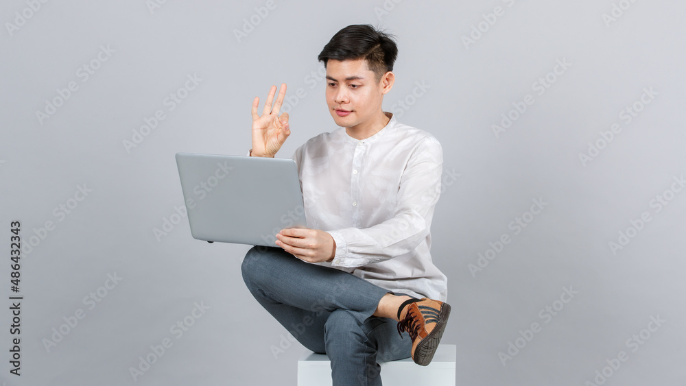 Studio shot of millennial Asian handsome young male model in casual outfit sitting on white square stool showing okay hand sign signal on video call with laptop notebook computer on gray background Studio shot of millennial Asian handsome young male model in casual outfit sitting on white square stool showing okay hand sign signal on video call with laptop notebook computer on gray background
