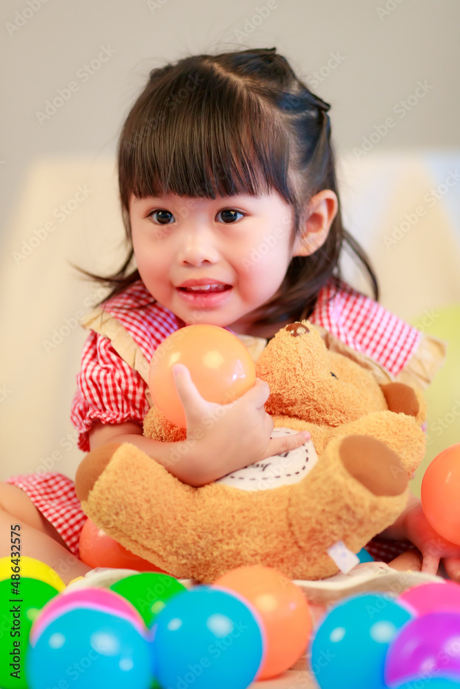 Portrait closeup studio shot of little cute kindergarten preschooler girl daughter in red dress sitting smiling hugging teddy bear doll on floor playing with colorful plastic balls on gray background