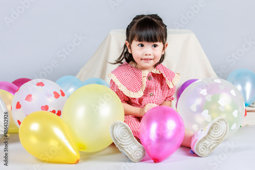 Portrait studio shot of little cute kindergarten preschooler kid girl daughter in red long dress sitting smiling alone on floor playing with colorful helium air party balloons on gray background