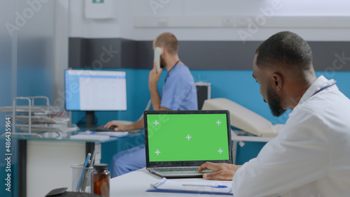 African american therapist doctor sitting at desk analyzing sickness report developing healthcare treatment in hospital office. Mock up green screen chroma key laptop computer with isolated display