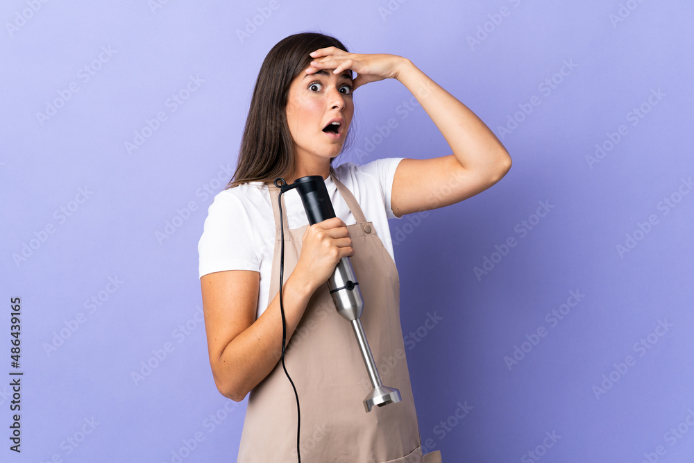 Brazilian woman using hand blender isolated on purple background doing surprise gesture while looking to the side