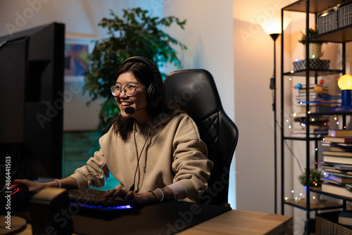 Smiling girl in sweatshirt and glasses with headphones plays video games online with friends talking to them through microphone, pass levels, rounds, strategy, victory, gamer room led neon lighting