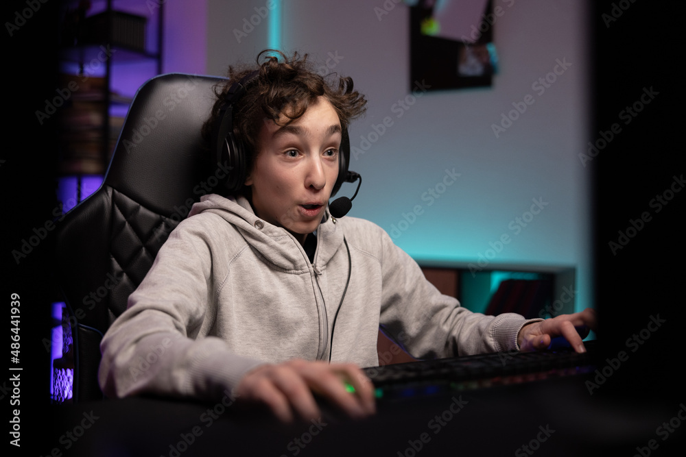 Surprised school-age gamer with headset boy sits late at night in front of computer monitor playing multiplayer shooter video games with team excitement of virtual world room lit up with led lights Surprised school-age gamer with headset boy sits late at night in front of computer monitor playing multiplayer shooter video games with team excitement of virtual world room lit up with led lights