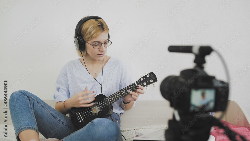 A young woman with glasses is sitting on the sofa, smiling and playing the ukulele to the camera. Playing the ukulele. Video Blogger A young woman with glasses is sitting on the sofa, smiling and playing the ukulele to the camera. Playing the ukulele. Video Blogger