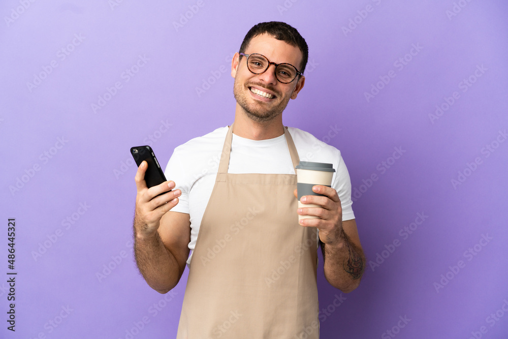 Brazilian restaurant waiter over isolated purple background holding coffee to take away and a mobile Brazilian restaurant waiter over isolated purple background holding coffee to take away and a mobile