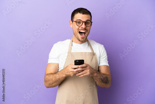 Brazilian restaurant waiter over isolated purple background surprised and sending a message