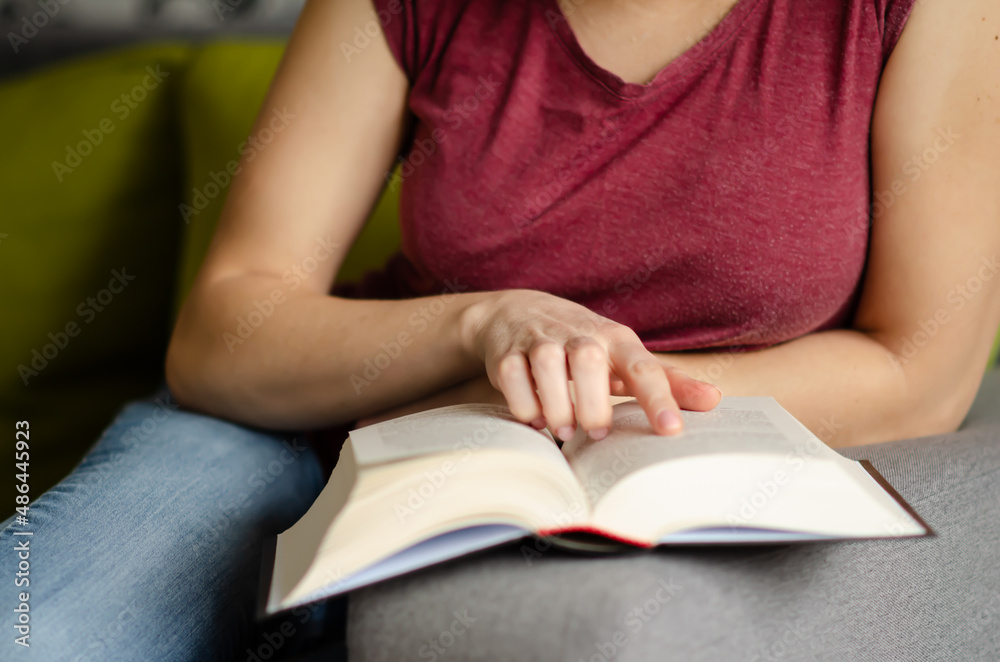 Selective focus on book read by caucasian woman in burgundy T-shirt and jeans sitting on gray sofa with pillows. Concept of rest in cozy house. Time alone with yourself. Selective focus on book read by caucasian woman in burgundy T-shirt and jeans sitting on gray sofa with pillows. Concept of rest in cozy house. Time alone with yourself.