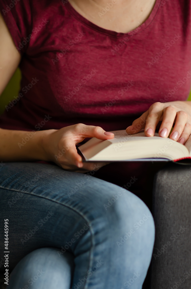 Fototapeta premium Selective focus on book read by caucasian woman in burgundy T-shirt and jeans sitting on gray sofa with pillows. Concept of rest in cozy house. Time alone with yourself.