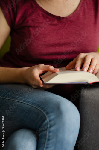 Selective focus on book read by caucasian woman in burgundy T-shirt and jeans sitting on gray sofa with pillows. Concept of rest in cozy house. Time alone with yourself.