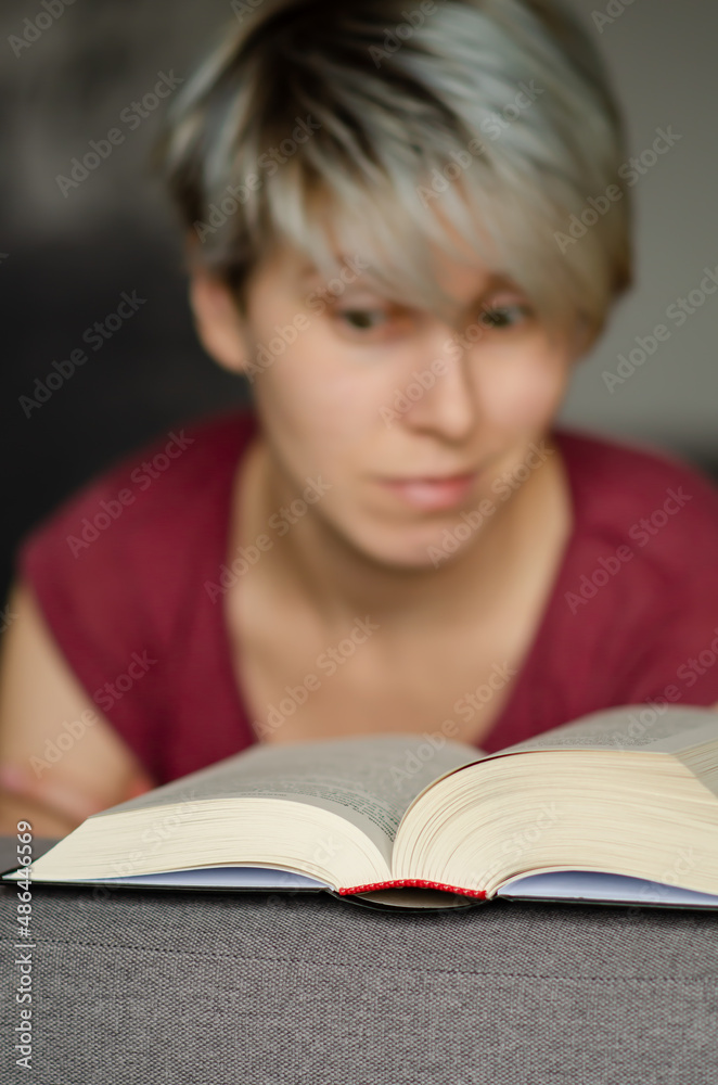 Selective focus on book read by smiling caucasian woman with short haircut on gray sofa. Concept of time alone with yourself. Young woman reads an interesting book and smiling. Selective focus on book read by smiling caucasian woman with short haircut on gray sofa. Concept of time alone with yourself. Young woman reads an interesting book and smiling.