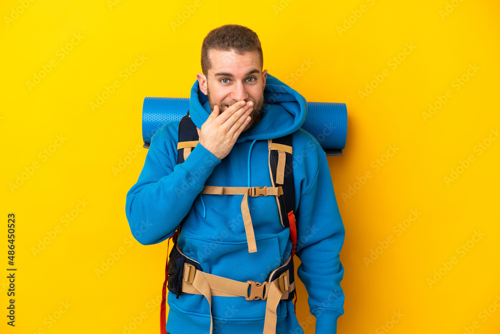 Young caucasian mountaineer man with a big backpack isolated on yellow background happy and smiling covering mouth with hand Young caucasian mountaineer man with a big backpack isolated on yellow background happy and smiling covering mouth with hand