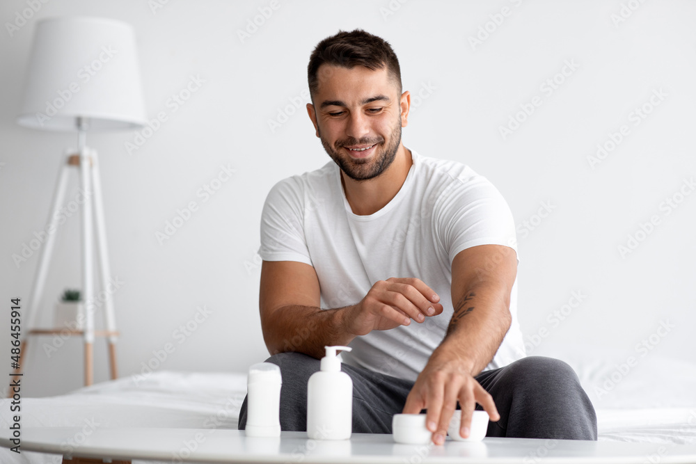 Smiling young caucasian muscular handsome man at table with jars of different moisturizing anti-aging cosmetics
