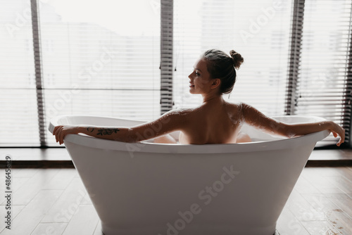 Back view of young lady taking foamy bath, looking aside at free space at home or hotel bathroom