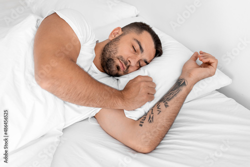 Tired peaceful calm millennial caucasian muscular male with closed eyes lies on white bed and sleeps in bedroom #486456525