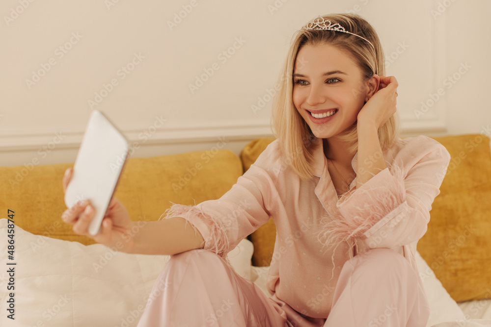 Beautiful fair-skinned young girl communicates by video communication through modern gadget while sitting in room. Pretty blonde woman wears diadem, pajamas. Technological device and emotion concept Beautiful fair-skinned young girl communicates by video communication through modern gadget while sitting in room. Pretty blonde woman wears diadem, pajamas. Technological device and emotion concept
