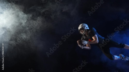 American football player in uniform, pads and helmet, catching pass and diving to ground with ball. Determination quarterback plays American football in dark arena among smoke and lights. Slow motion. photo