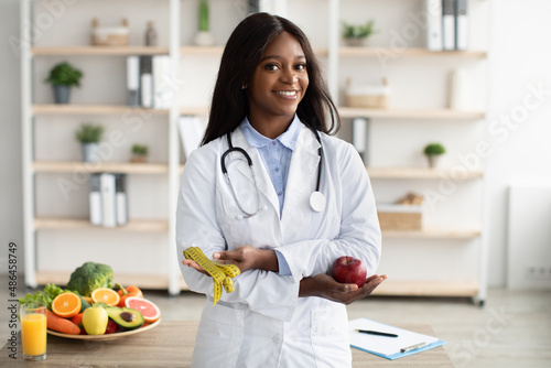 Portrait of cheerful young black female dietician with apple and tape recommending fruits and vegetables, free space