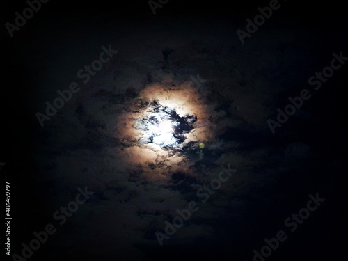 The moon illuminates the sky around it at night, through the clouds and darkness 