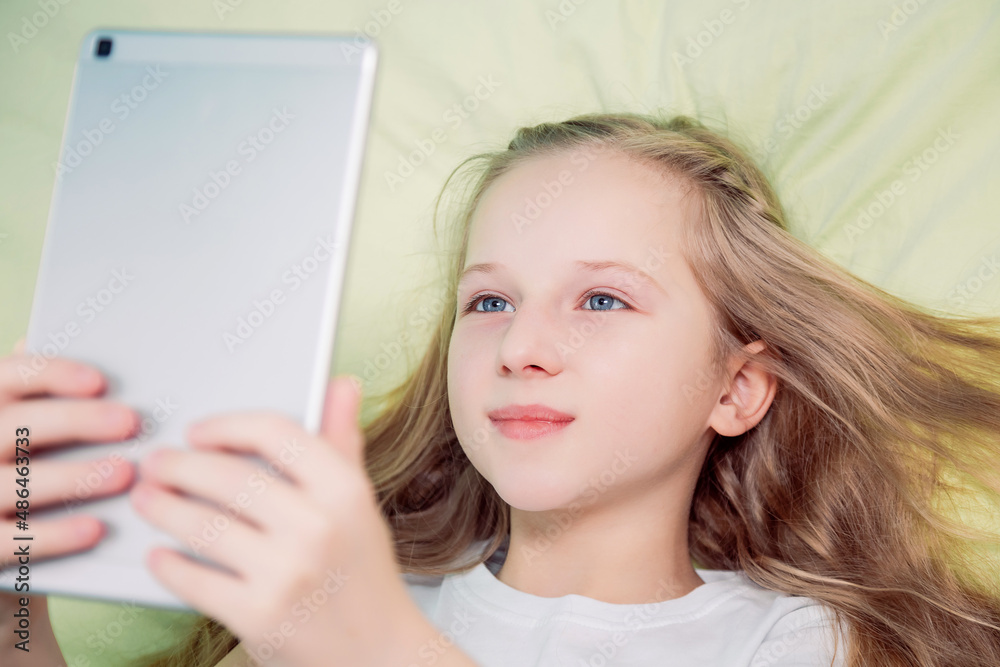 Naklejka premium Beautiful girl lying on the bed looks into the tablet. The girl takes a selfie.