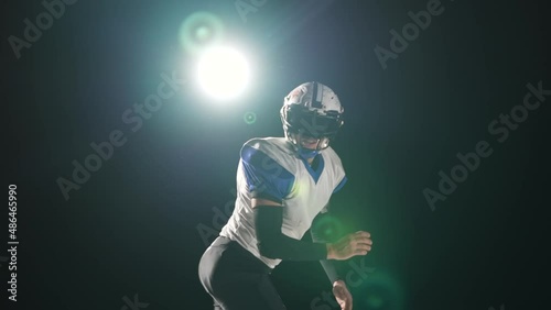 American football player wearing uniform, pads and helmet catches pass and runs on attack against black background with spotlight. Players run attacking to score touchdown points. Slow motion. photo