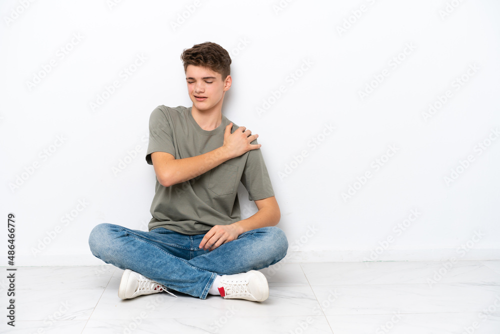 Teenager Russian man sitting on the floor isolated on white background suffering from pain in shoulder for having made an effort