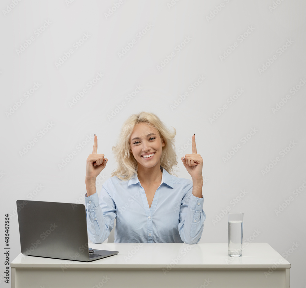Happy businesswoman sitting at desk with laptop and pointing fingers up at copy space on light studio background, crop Happy businesswoman sitting at desk with laptop and pointing fingers up at copy space on light studio background, crop