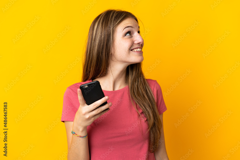Young woman using mobile phone isolated on yellow background looking up while smiling Young woman using mobile phone isolated on yellow background looking up while smiling