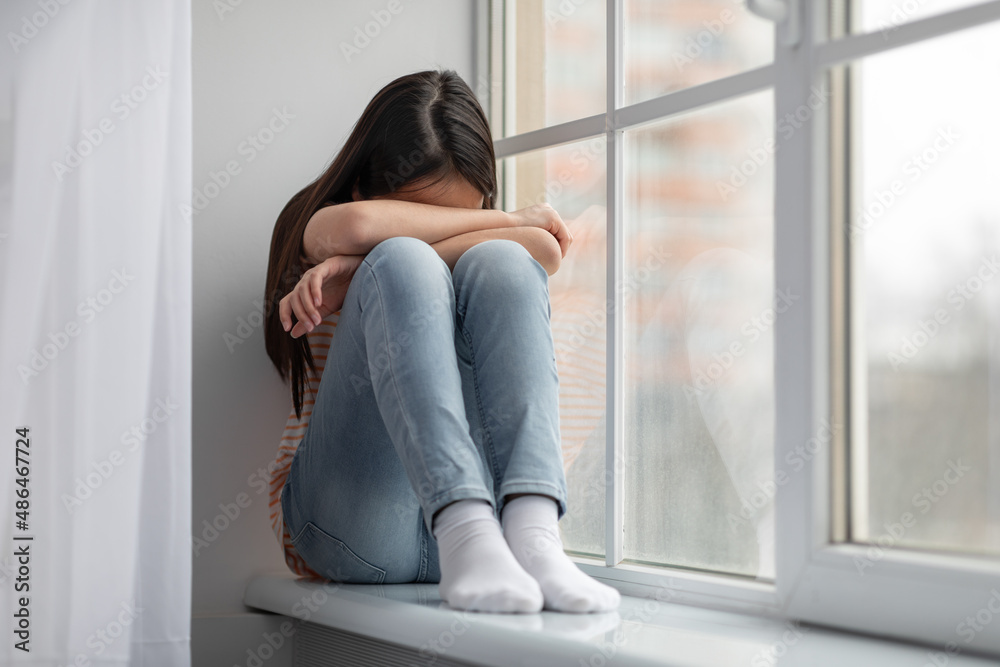 Upset asian girl schooler sitting on windowsill and crying