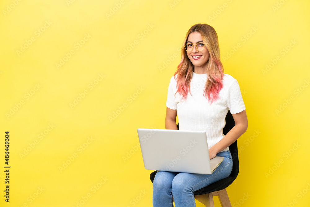 Naklejka premium Young woman sitting on a chair with laptop over isolated yellow background posing with arms at hip and smiling