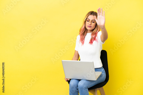Young woman sitting on a chair with laptop over isolated yellow background making stop gesture and disappointed