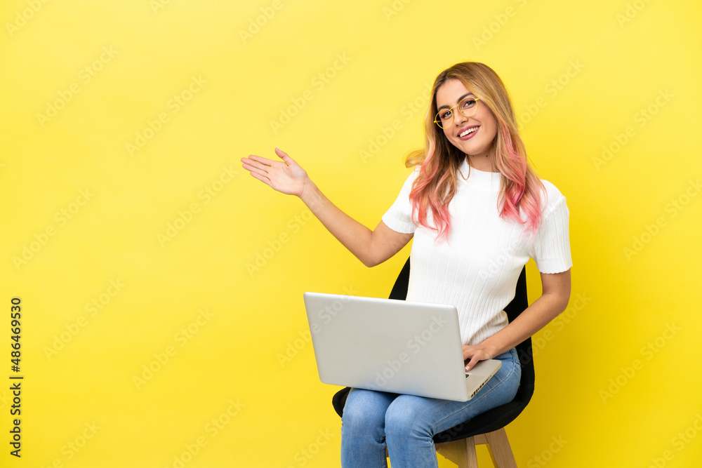 Young woman sitting on a chair with laptop over isolated yellow background extending hands to the side for inviting to come Young woman sitting on a chair with laptop over isolated yellow background extending hands to the side for inviting to come