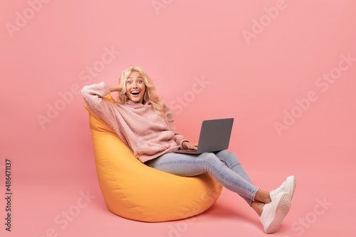 Excited young lady sitting in beanbag chair with laptop, shouting wow, celebrating great news, pink background photo