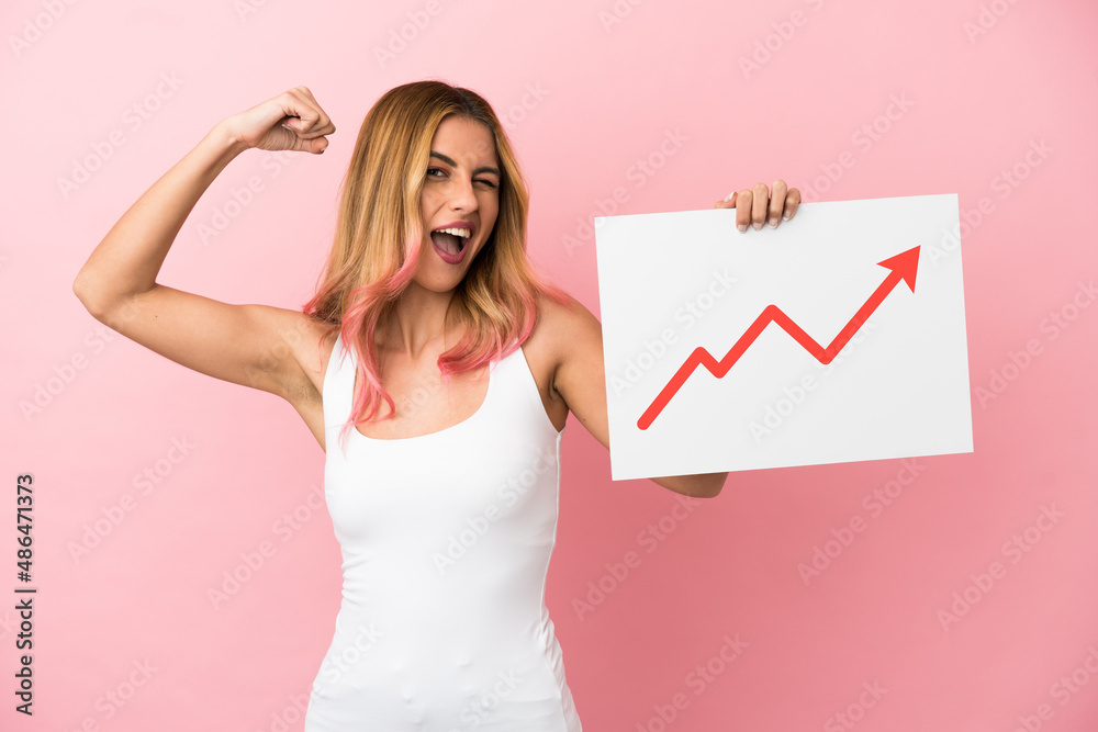 Young woman over isolated pink background holding a sign with a growing statistics arrow symbol and doing strong gesture Young woman over isolated pink background holding a sign with a growing statistics arrow symbol and doing strong gesture