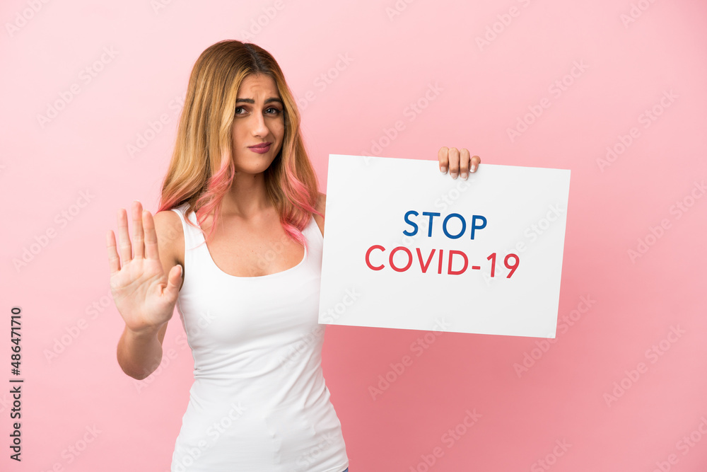 Young woman over isolated pink background holding a placard with text Stop Covid 19 and doing stop sign Young woman over isolated pink background holding a placard with text Stop Covid 19 and doing stop sign