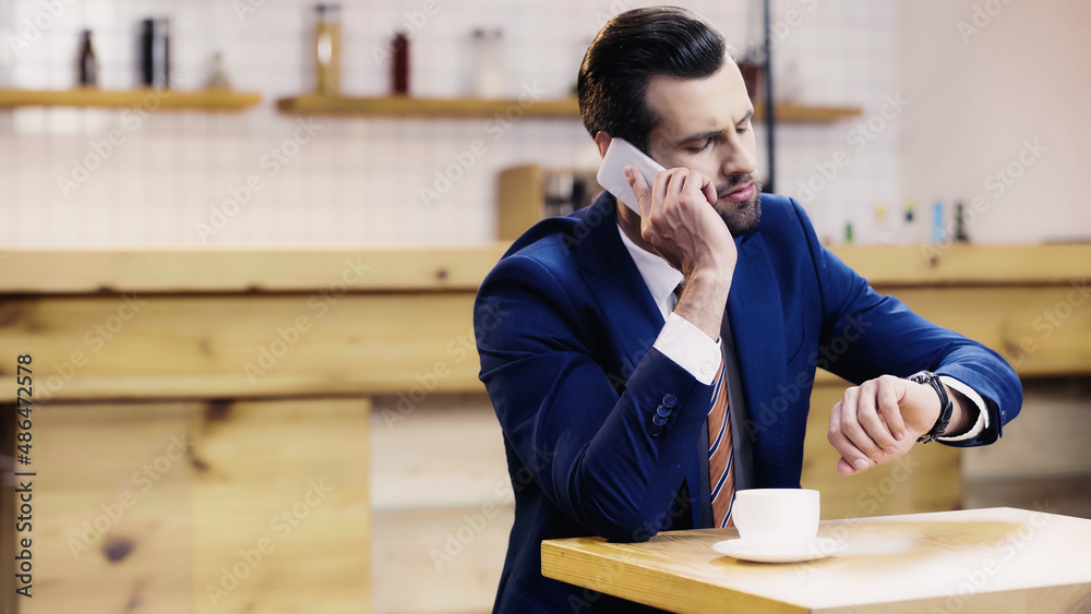 bearded businessman in suit talking on smartphone and checking time in cafe